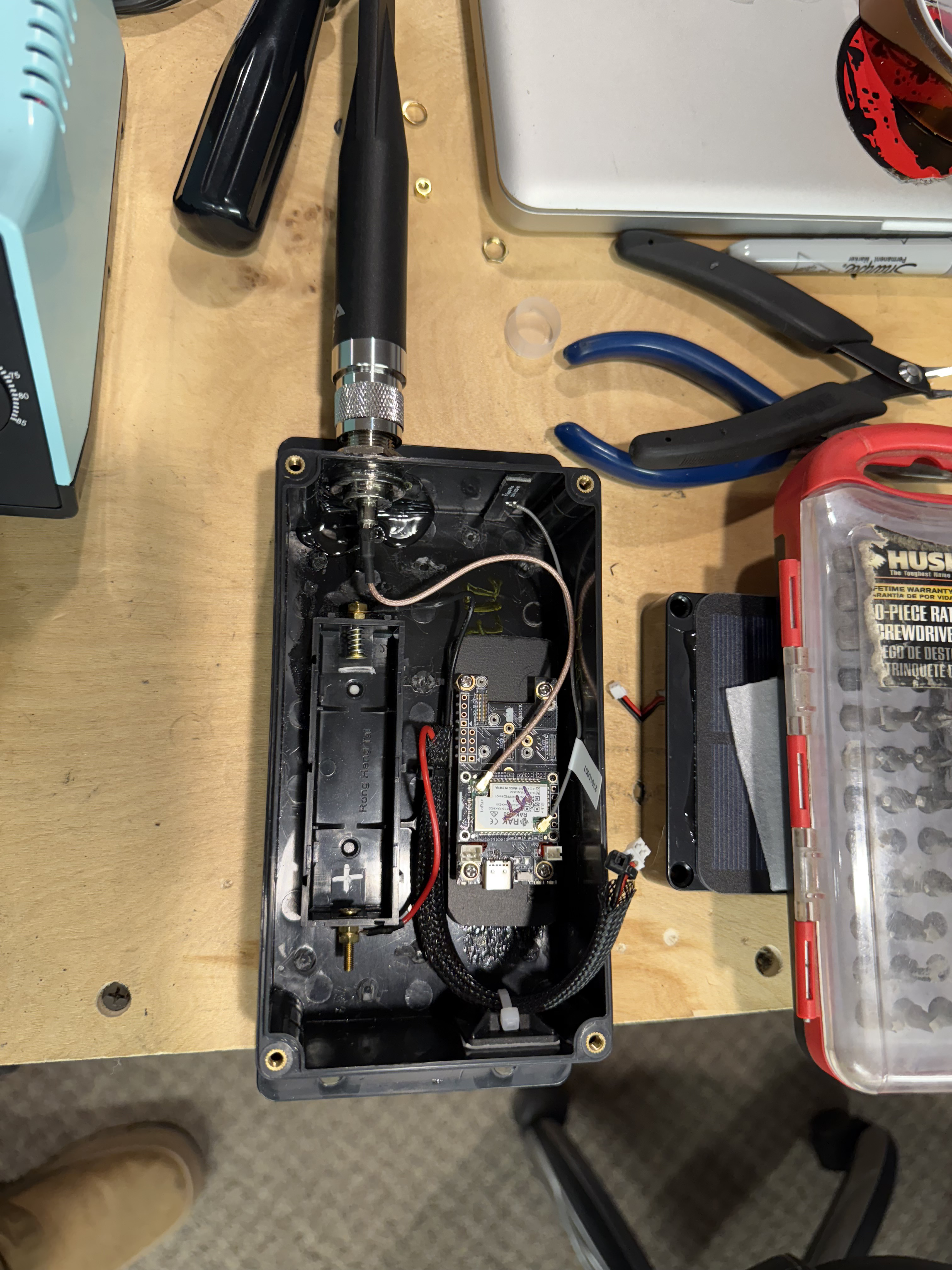 Open black weatherproof enclosure with RAK radio module, battery holder, and sealed antenna bulkhead on a workbench