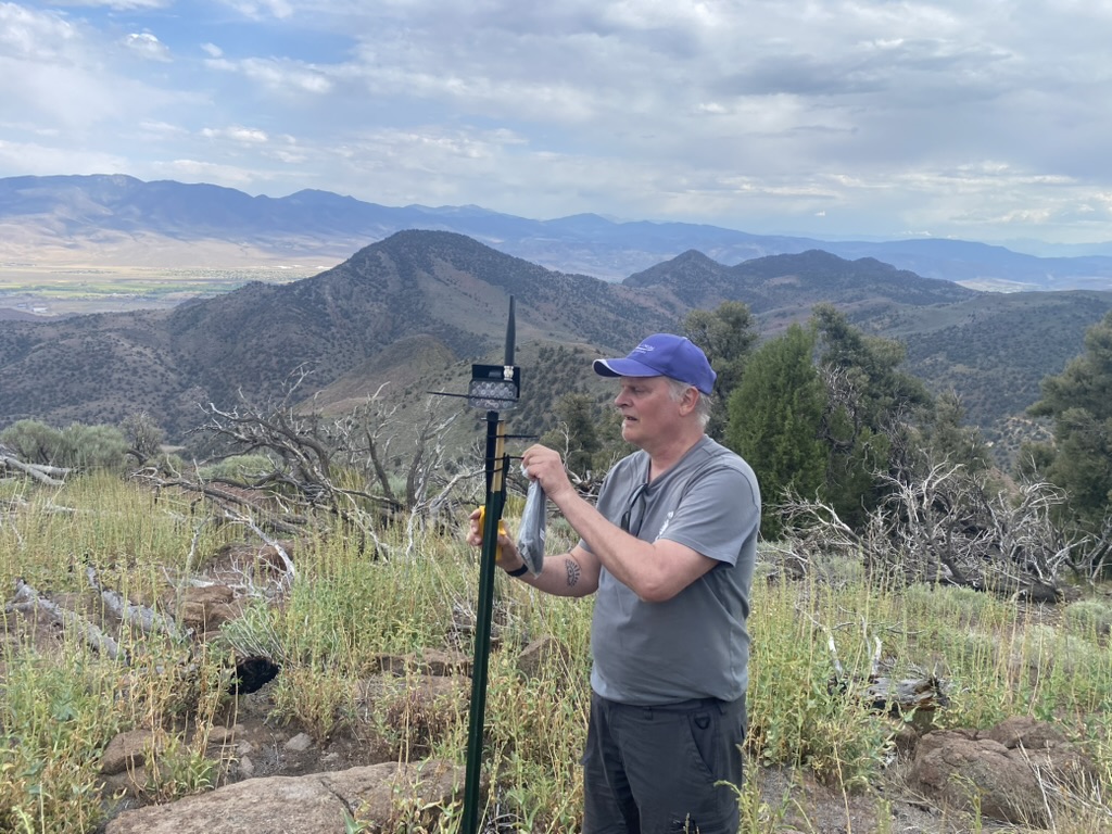 Tim driving a T-post mesh node into the rocky summit while holding a battery pack