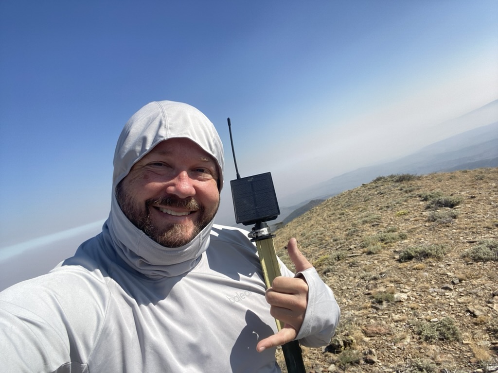 Ben holding a mesh repeater on a T-post at the Fox Mountain summit, wildfire smoke haze in the valley below