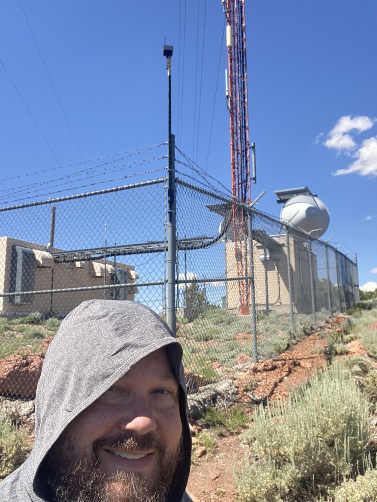 Ben in a sun hoodie at the High Sierra Communications compound on McClellan Peak, T-stake node mount on the fence behind him