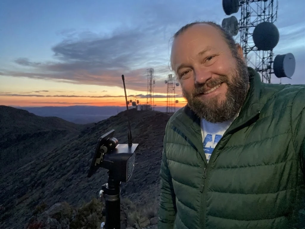 Ben with a MeshCore node on Toulon Peak at dusk, microwave tower and sunset behind him